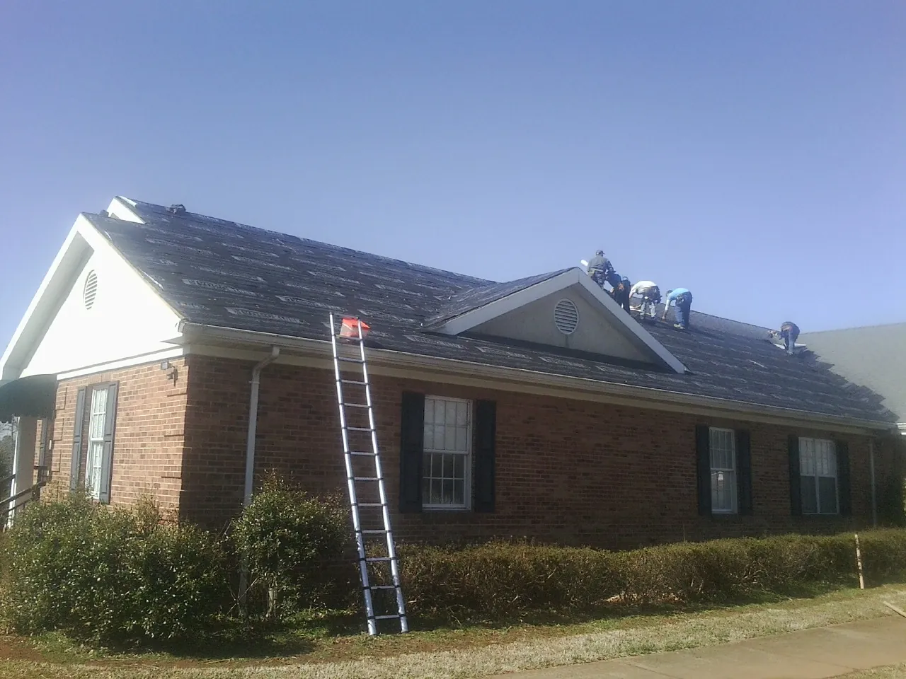 Crew tearing shingles off a brick ranch roof with a ladder set — Emergency Roof Repair work by our crew in Monument Beach