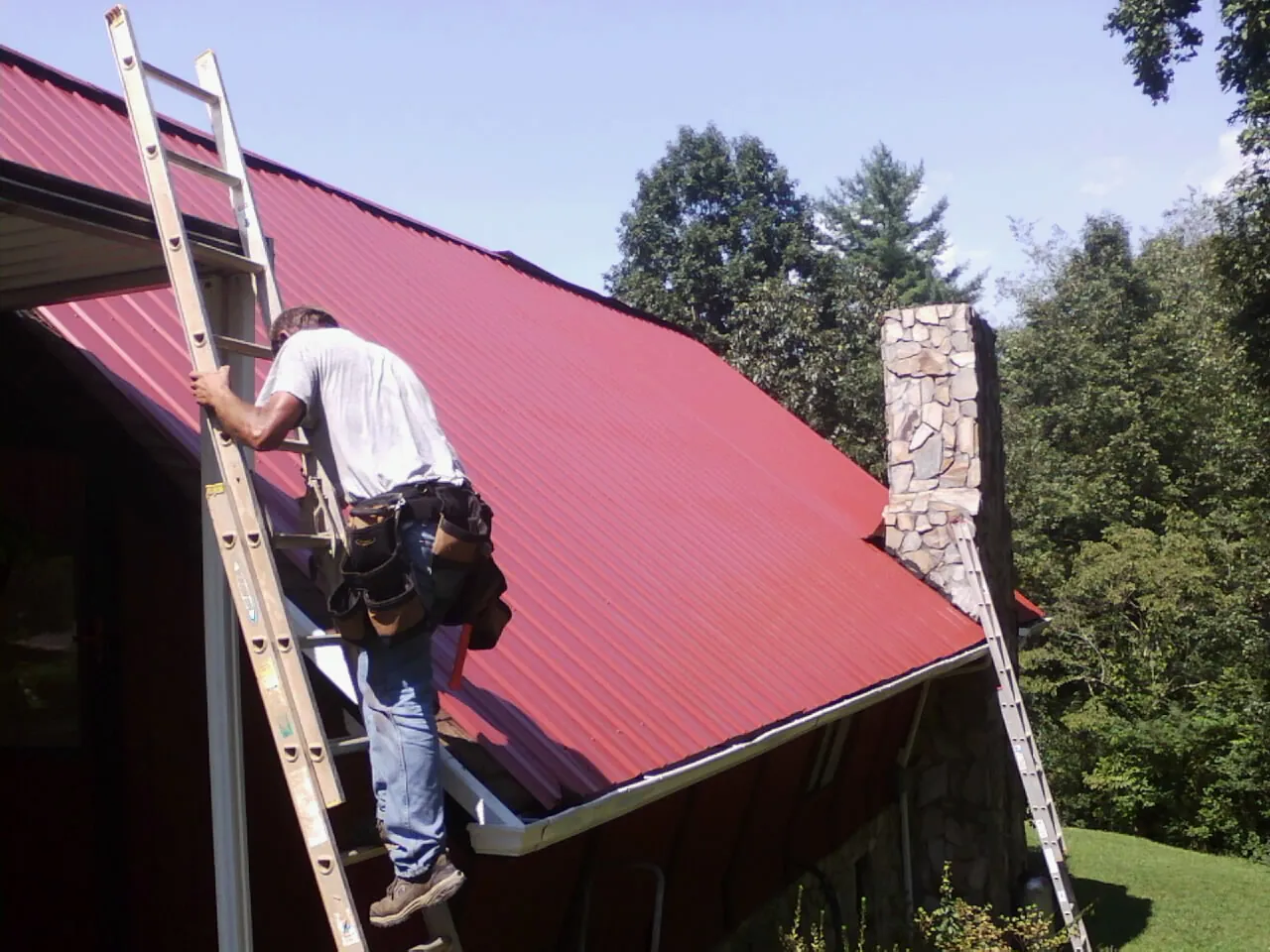 Roofer on a ladder beside a red metal roof and stone chimney — Soffit & Fascia Repair by Roofing Brothers in Monument Beach
